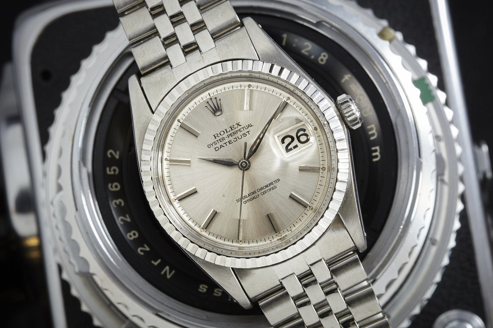 A close-up of a Rolex Datejust by Rolex with Alpha hands, fluted bezel, jubilee bracelet, and date showing 26, resting on a vintage camera lens.