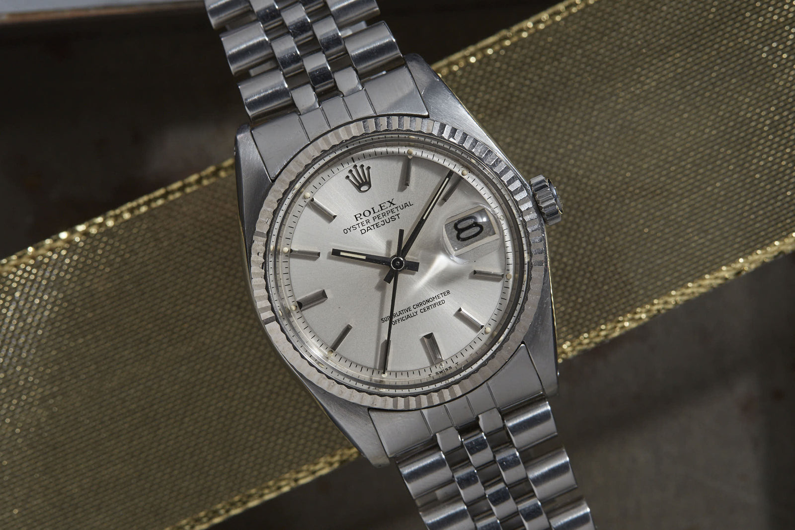 A Rolex Datejust by Rolex, featuring a silver dial, date display, fluted bezel, and metallic link bracelet, is elegantly displayed against a textured gold ribbon background.