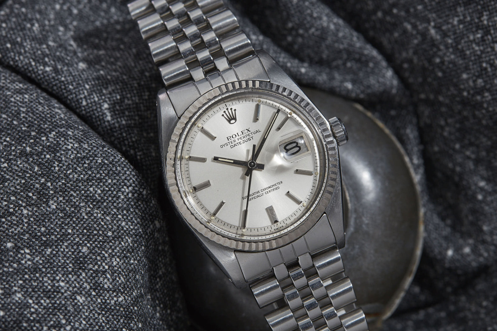 A Rolex Datejust wristwatch with a metal link band and white dial showing the date, featuring the iconic Rolex Oyster case, rests on a textured dark gray fabric background.