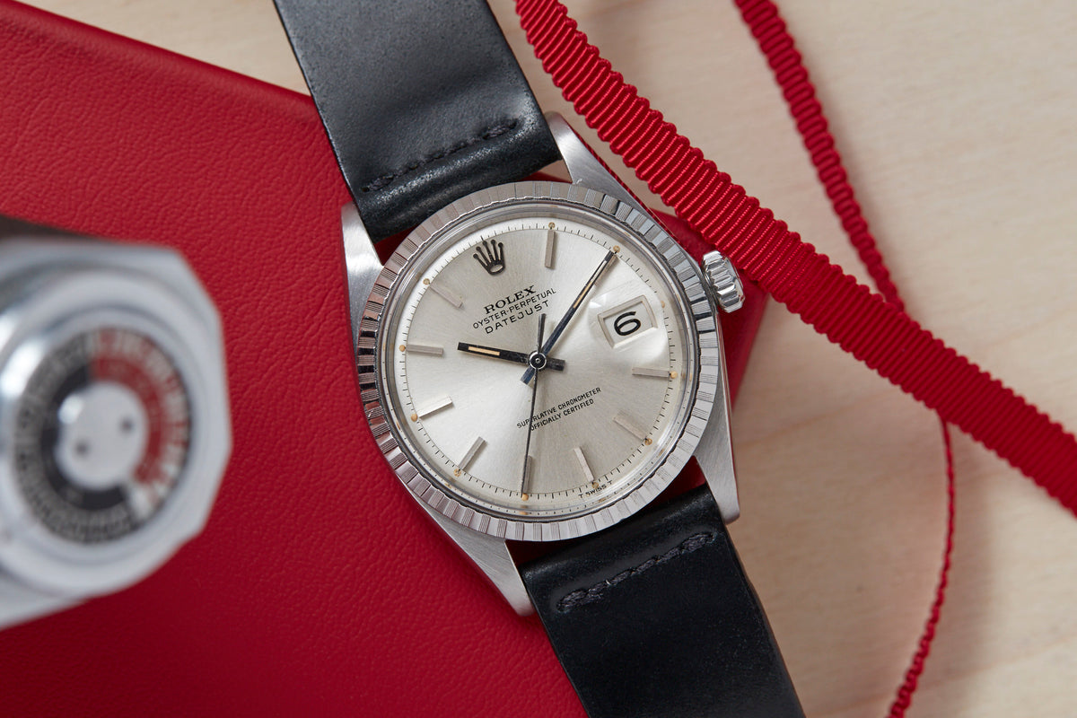 Stainless steel watch with silver dial, date window, and black leather strap on red fabric background.
