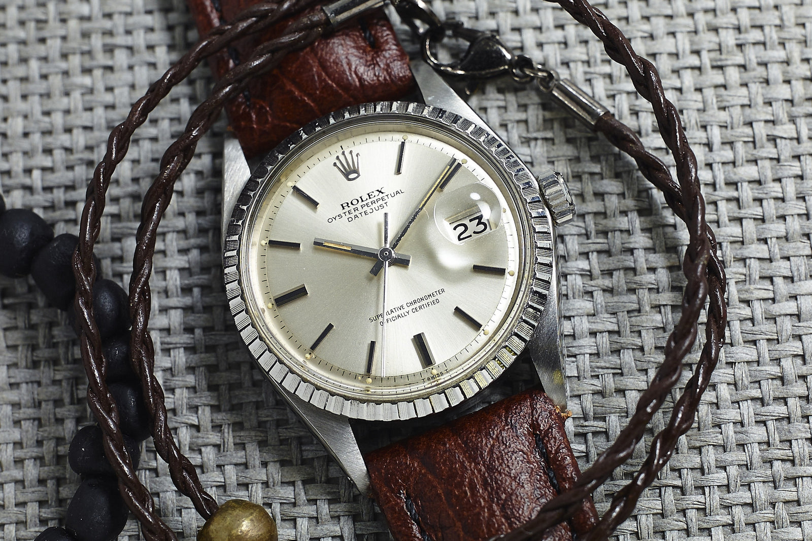 A close-up of a Rolex Datejust 1603 Silver Dial - 1970 with a brown leather strap and date set to the 23rd, displayed on a woven gray surface beside braided leather and beaded bracelets.