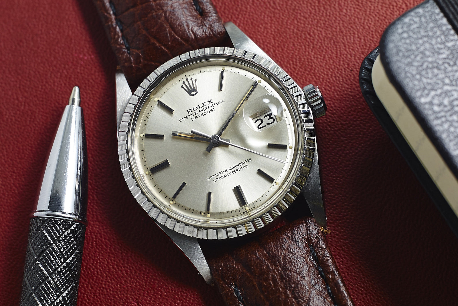 A close-up of a Rolex Datejust 1603 Silver Dial - 1970 wristwatch with a brown leather strap shows the date as 23. The watch rests on a red surface beside a silver pen and part of a black item.