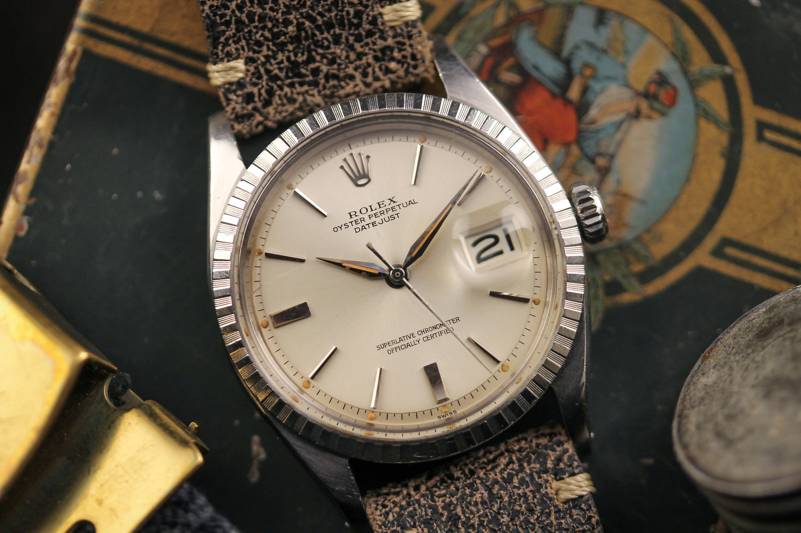 A close-up of a Rolex Datejust 1603 Sword Hands-1953 wristwatch by Rolex, featuring a cream dial, silver sword hands, date window at 3 o’clock, and a worn textured strap against a backdrop of colorful vintage items.