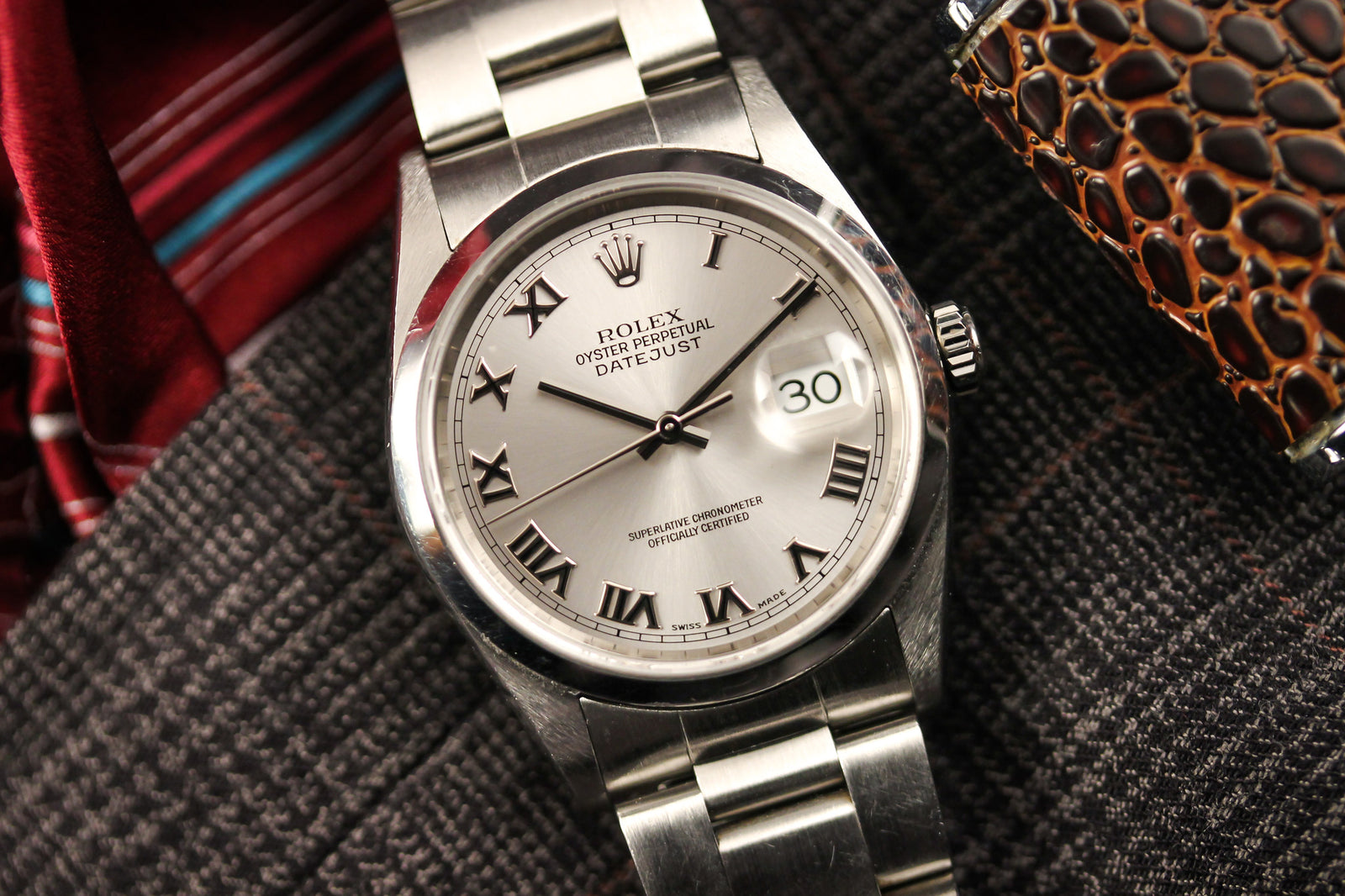 A Rolex Datejust 16200 with a silver sunburst dial, white Roman numerals, and date display by Rolex rests on textured fabric beside a patterned tie and brown accessory.