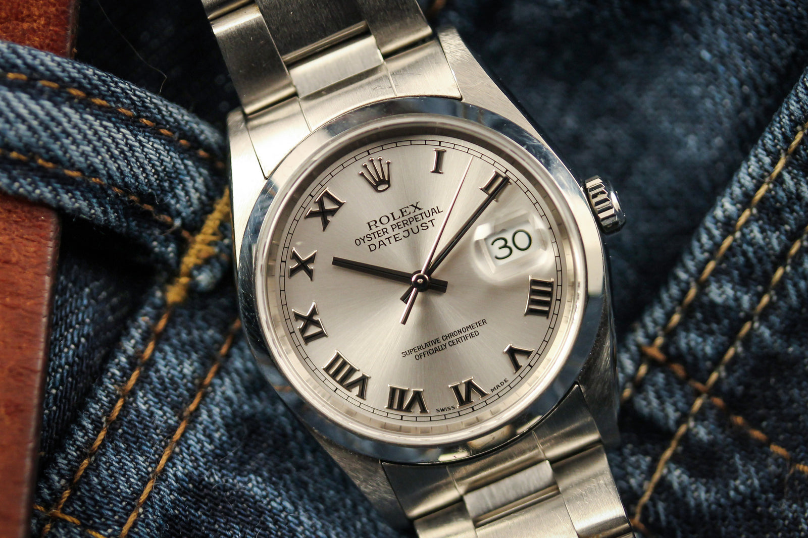 A Rolex Datejust 16200 by Rolex, featuring a silver sunburst dial with Roman numerals and metal bracelet, rests on blue denim fabric with the date window showing the 30th.