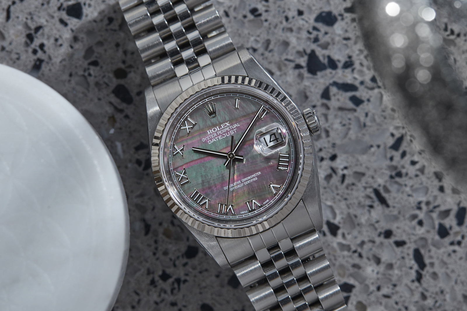 A Rolex Datejust Mother of Black Pearl Dial watch with Roman numerals is displayed on a speckled gray surface beside a round white object. Detailed description coming soon!.