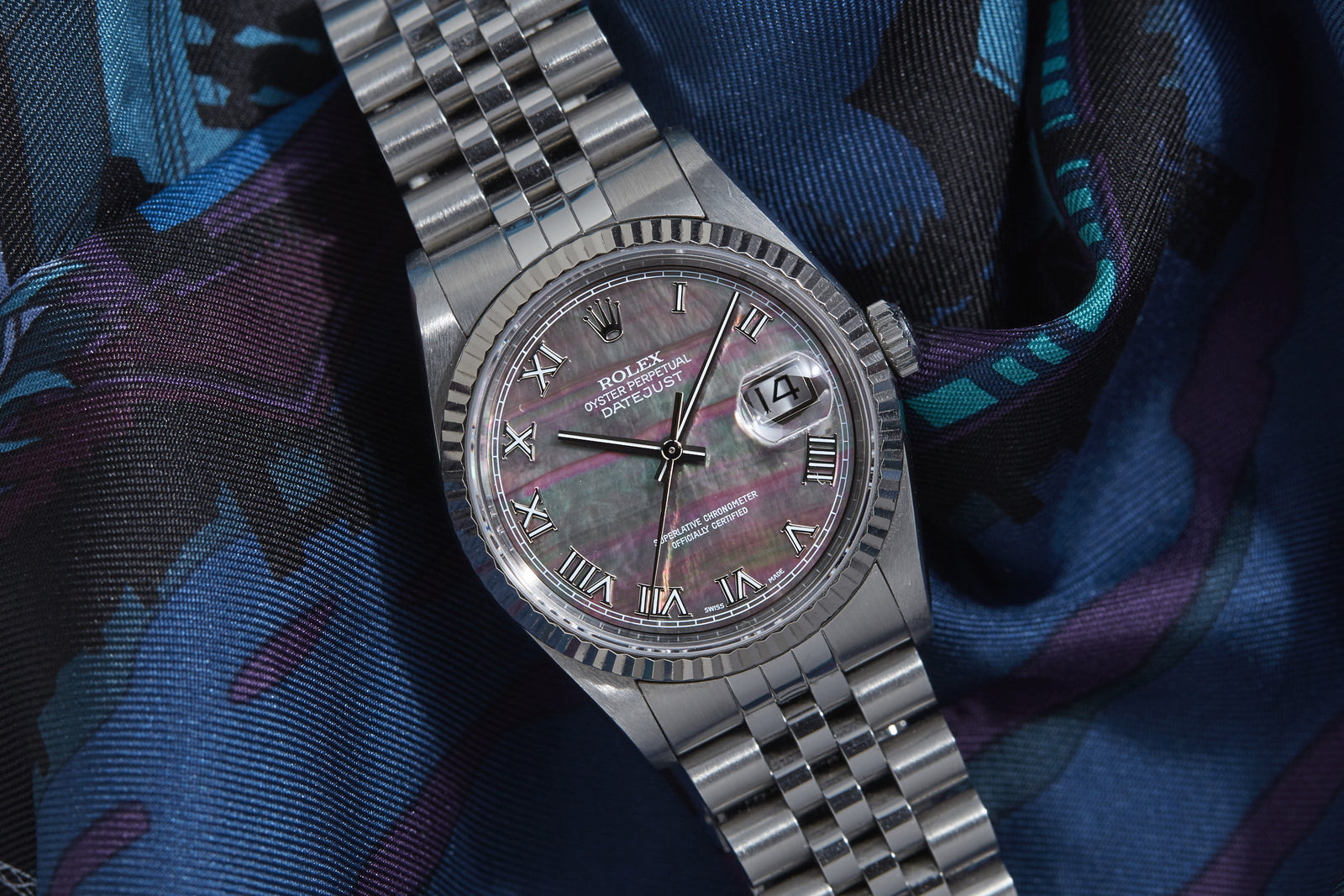 The Rolex Datejust Mother of Black Pearl Dial by Rolex features a silver case, black and purple mother-of-pearl dial with Roman numerals, showcased on a vibrant patterned fabric. Detailed description coming soon!.