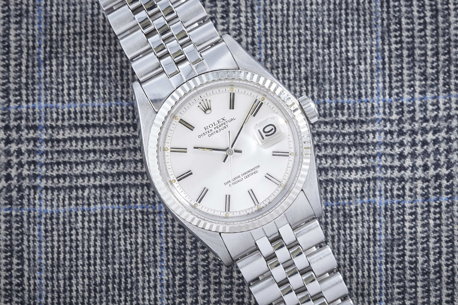 A Rolex Datejust by Rolex featuring a white dial and date window rests on gray plaid fabric with blue lines.