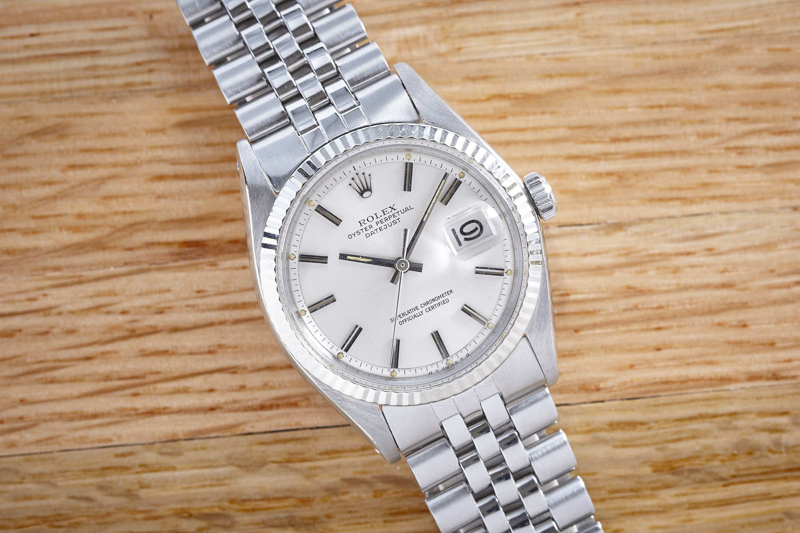 A Rolex Datejust wristwatch by Rolex, featuring a silver stainless steel band and white dial with date display, plus matching silver hands and hour markers, rests on a wooden surface.