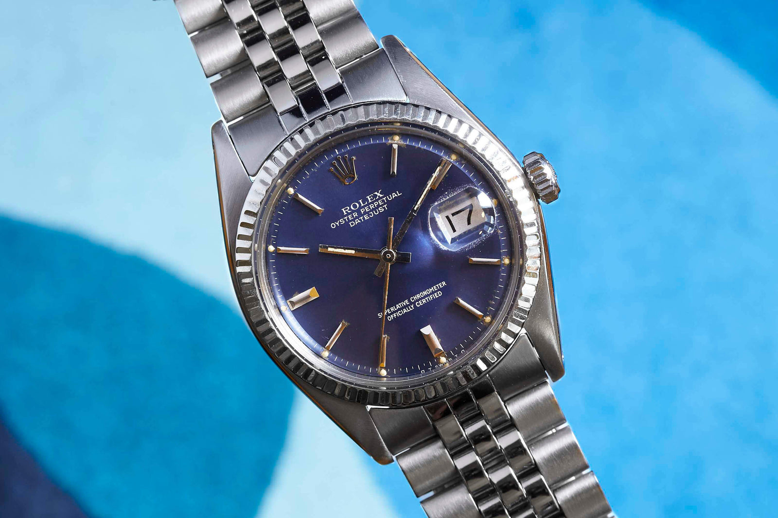 The Rolex Datejust by Rolex is shown with a blue dial, date display, silver hour markers and hands, and a metal link bracelet, set against a blue abstract background.