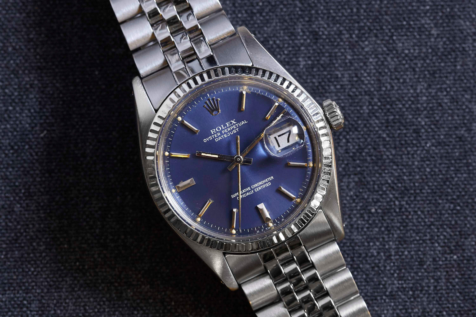 A Rolex Datejust wristwatch by Rolex features a blue dial, gold-toned hands and hour markers, date display, and metal link band, shown resting on a dark textured fabric background.