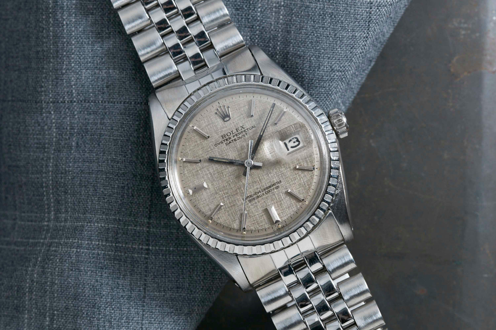 A Rolex Datejust 1601 Grey Linen wristwatch by Rolex, featuring a textured gray dial, automatic movement, date at 3 o’clock, and jubilee bracelet, displayed on folded gray fabric.