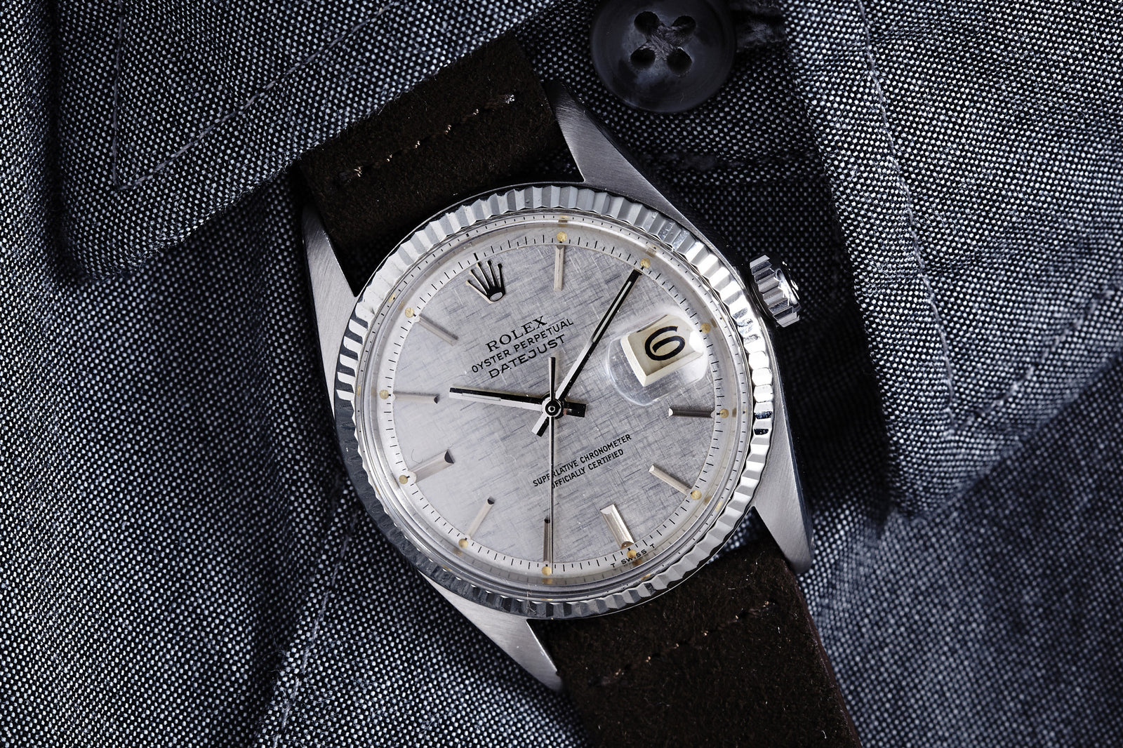 A close-up of a Rolex Datejust Reference 1601 Linen Dial - 1973 with a silver dial and date display, worn on a black leather strap over the cuff of a gray textured button-up shirt.