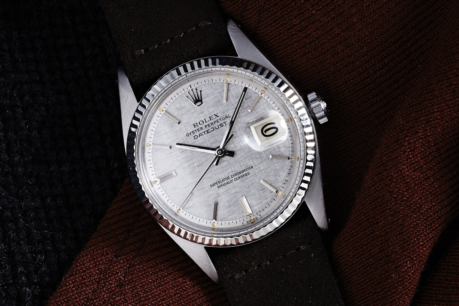 A Rolex Datejust Reference 1601 Linen Dial (1973) with a fluted bezel and date at 6 o’clock, rests on a brown and black textured fabric background.