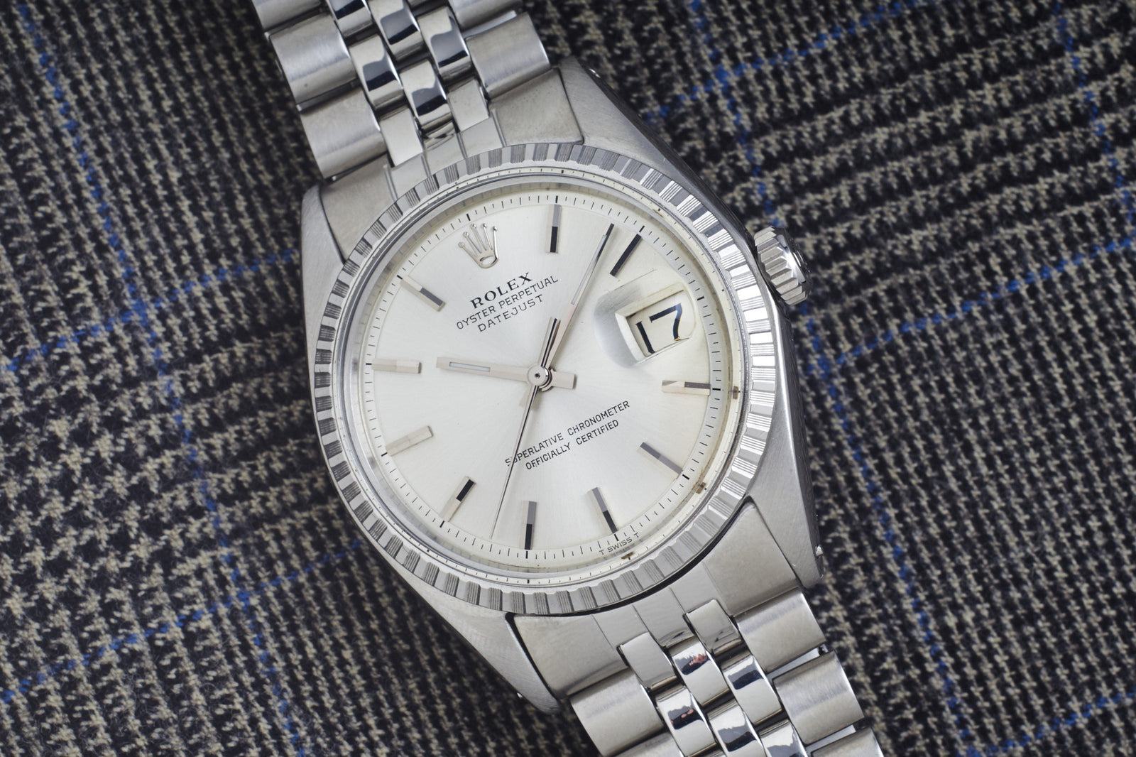 The Rolex Datejust Reference 1603 - 1966 by Rolex, featuring a silver case, white dial, date window, and stainless steel jubilee bracelet, is displayed on textured dark plaid fabric—embodying classic luxury.