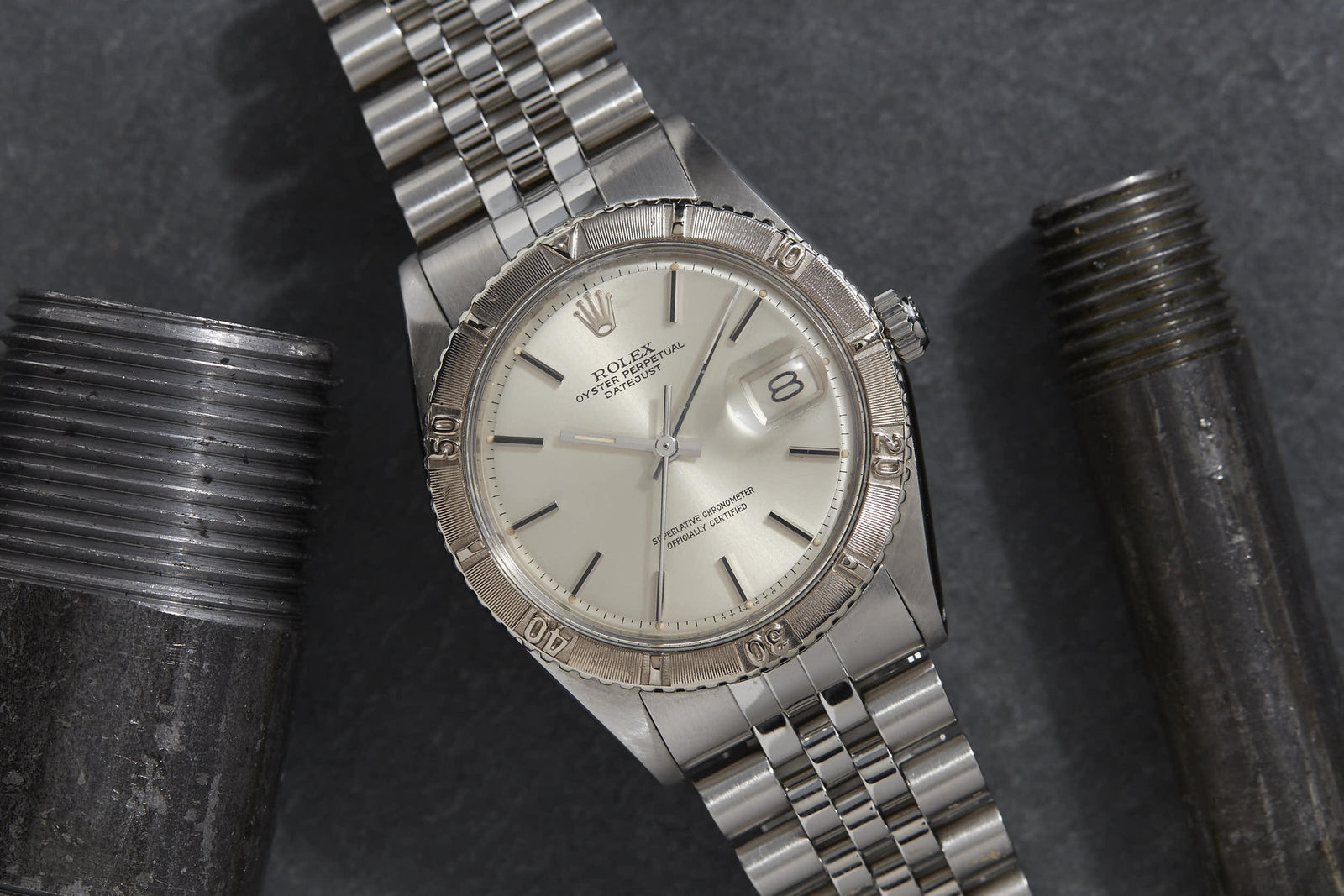 A Rolex Turn-O-Graph "Thunderbird" watch is positioned between two large metal bolts on a dark, textured surface, showcasing its timeless design and signature elegance.