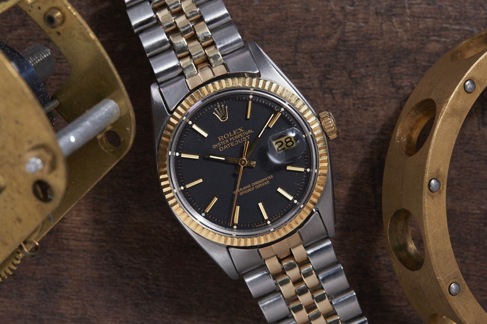 A Rolex Datejust by Rolex, featuring a black dial, gold hour markers, and a Jubilee bracelet, sits on a wooden surface amid brass mechanical parts.