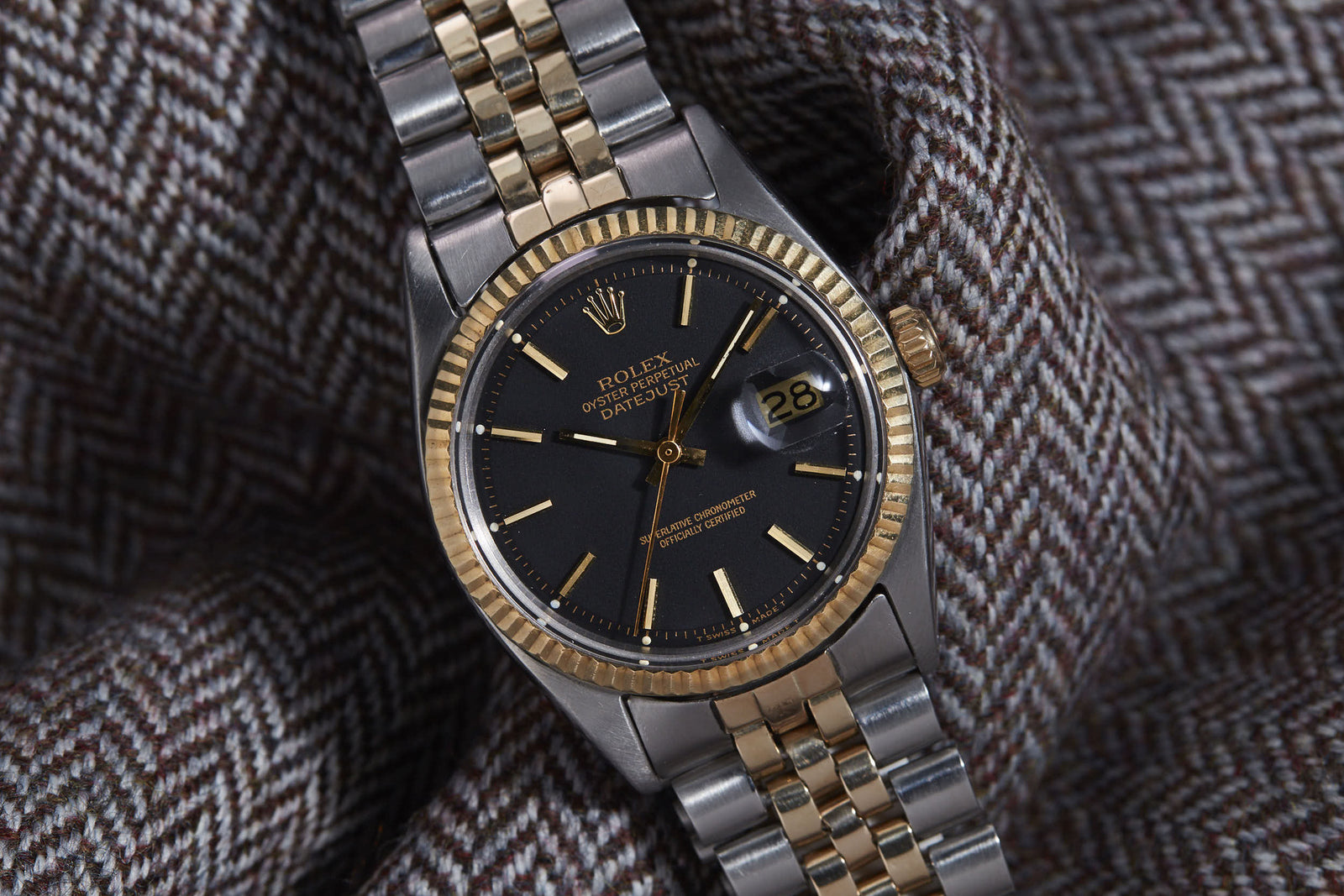 A Rolex Datejust watch with a black dial, gold bezel, and date window rests on brown and white herringbone fabric, showcasing the signature Jubilee bracelet for elegance.