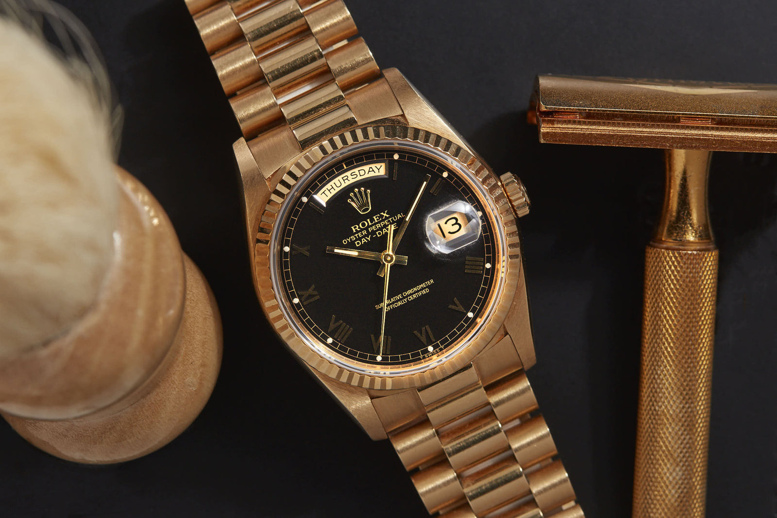 A Rolex Day-Date gold watch by Rolex with a black dial displaying day and date, set next to a gold razor and shaving brush on a dark surface.