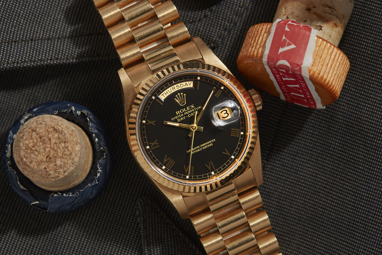 A gold Rolex Day-Date from Rolex with a black dial and Roman numerals rests on dark fabric, flanked by blue and red corks. This iconic watch displays Thursday and the 13th in signature Oyster Perpetual style.