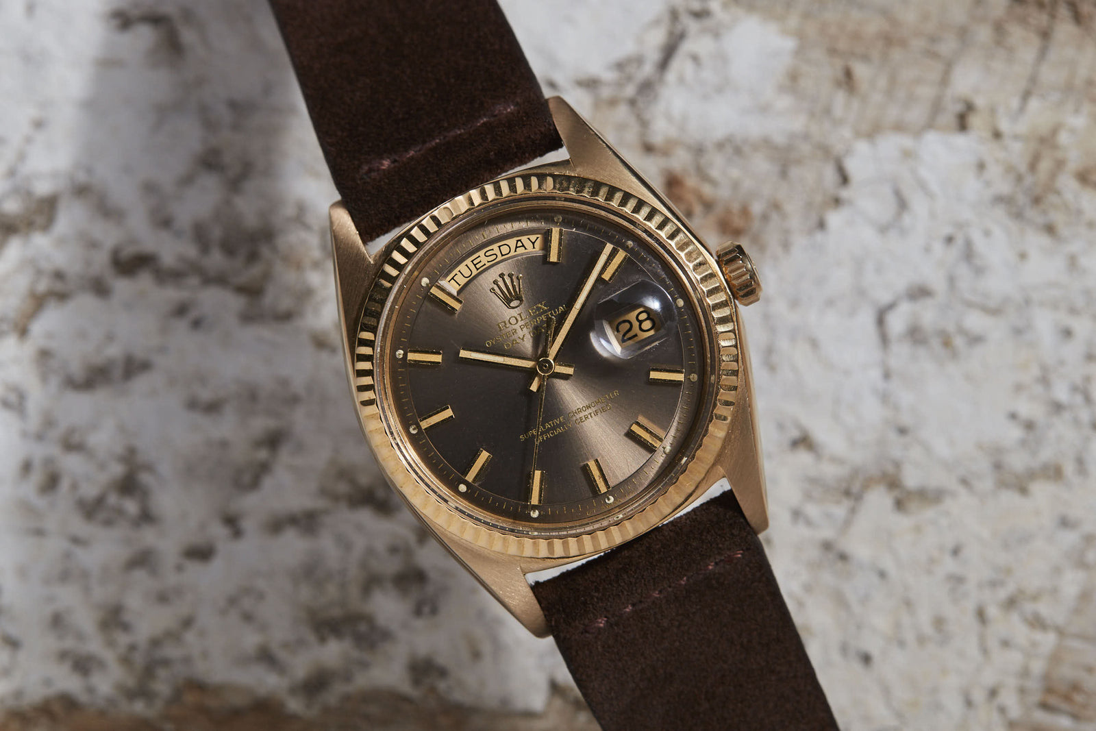 A Rolex Day-Date by Rolex, featuring a gold bezel, brown dial, day-date display, dark brown leather strap, and automatic movement, rests on a textured, weathered surface.
