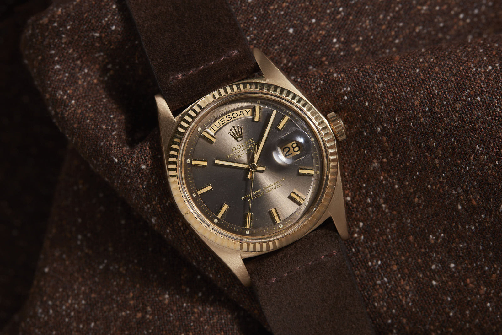 The Rolex Day-Date by Rolex features a dark brown dial, brown leather strap, and day-date display, elegantly placed on textured brown fabric.
