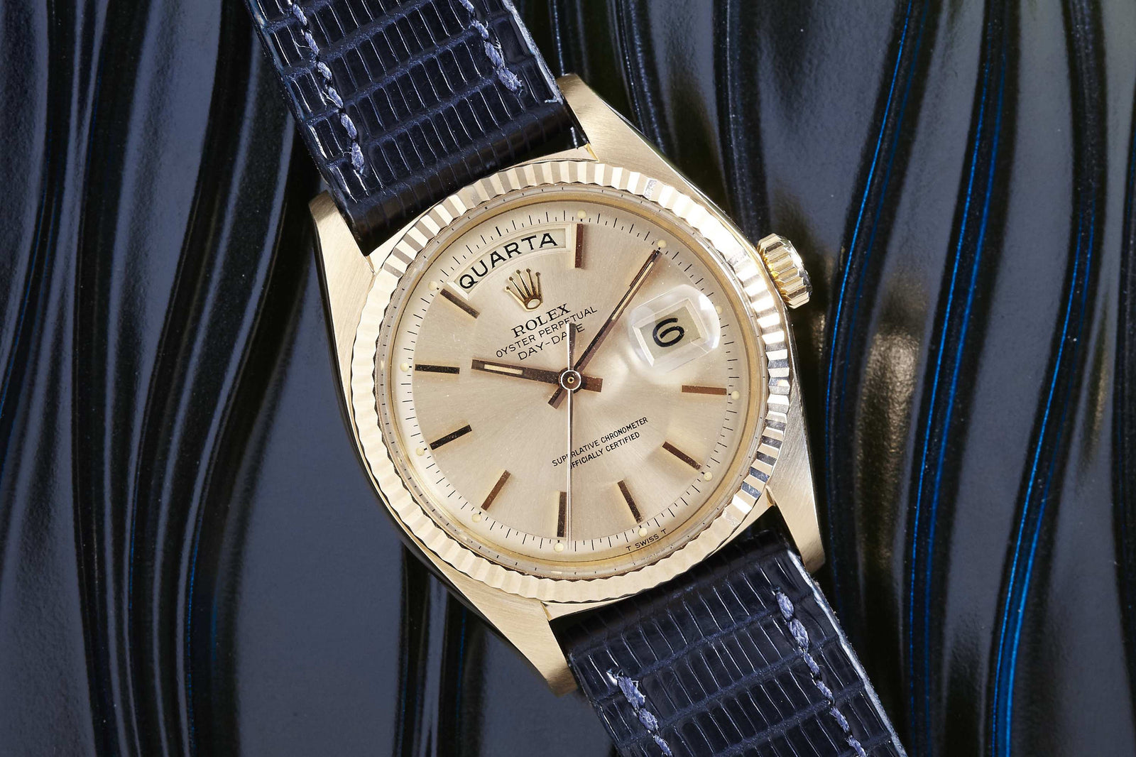 A Rolex Day-Date watch by Rolex with a beige dial, day and date displays, and a black textured strap rests on a glossy black wavy surface accented with blue.