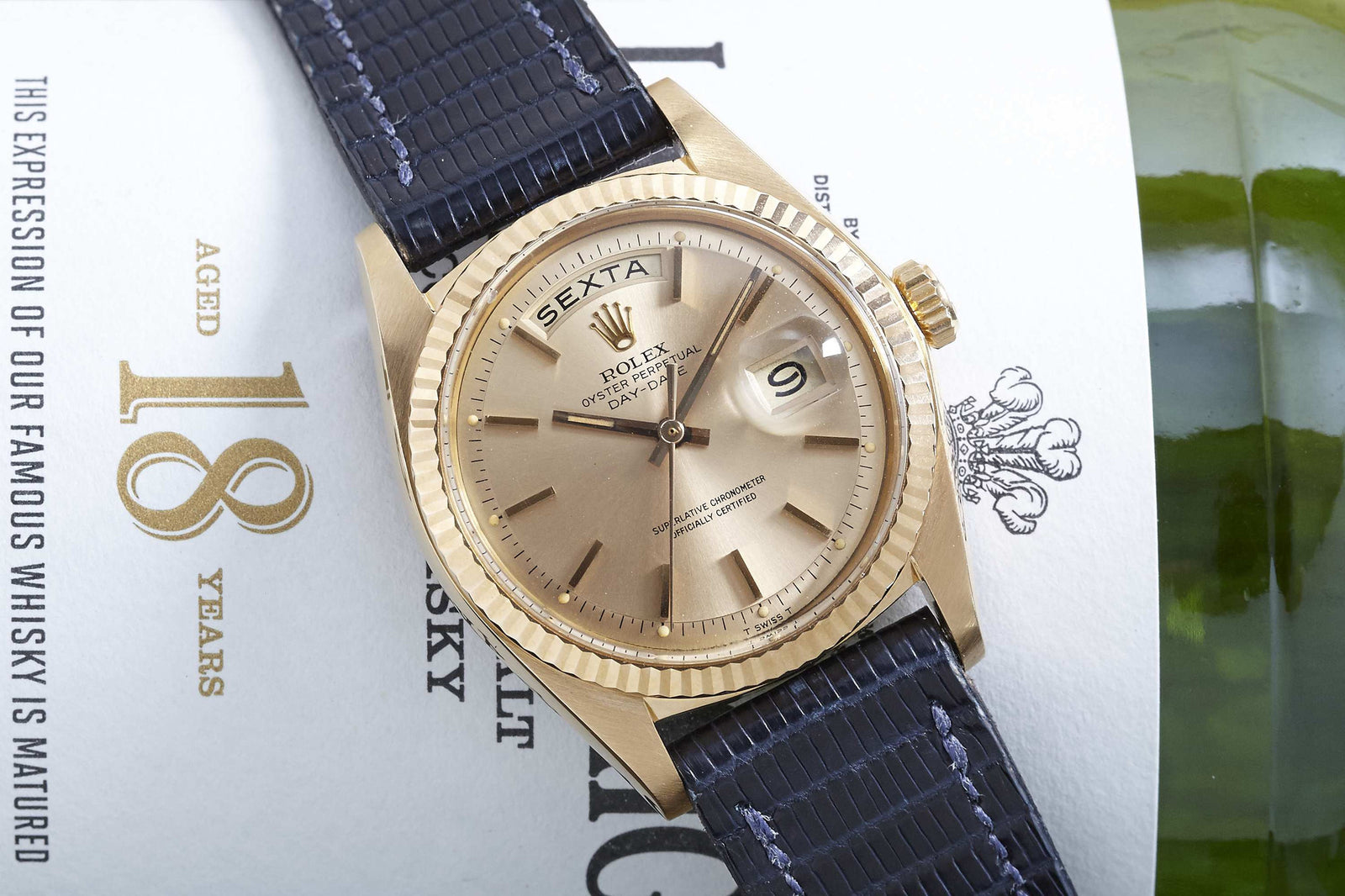 A Rolex Day-Date by Rolex in 18k gold with a black strap sits atop a whisky label marked “Aged 18 Years,” with part of a green bottle visible on the right.