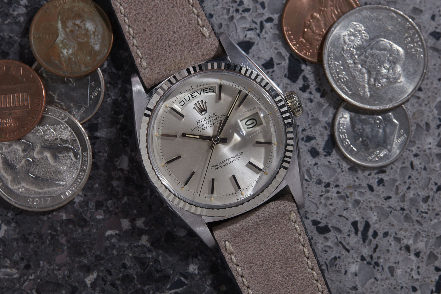 A Rolex Day Date by Rolex with a beige leather strap sits on a speckled gray surface, surrounded by various U.S. coins—quarters, nickels, dimes, and pennies.