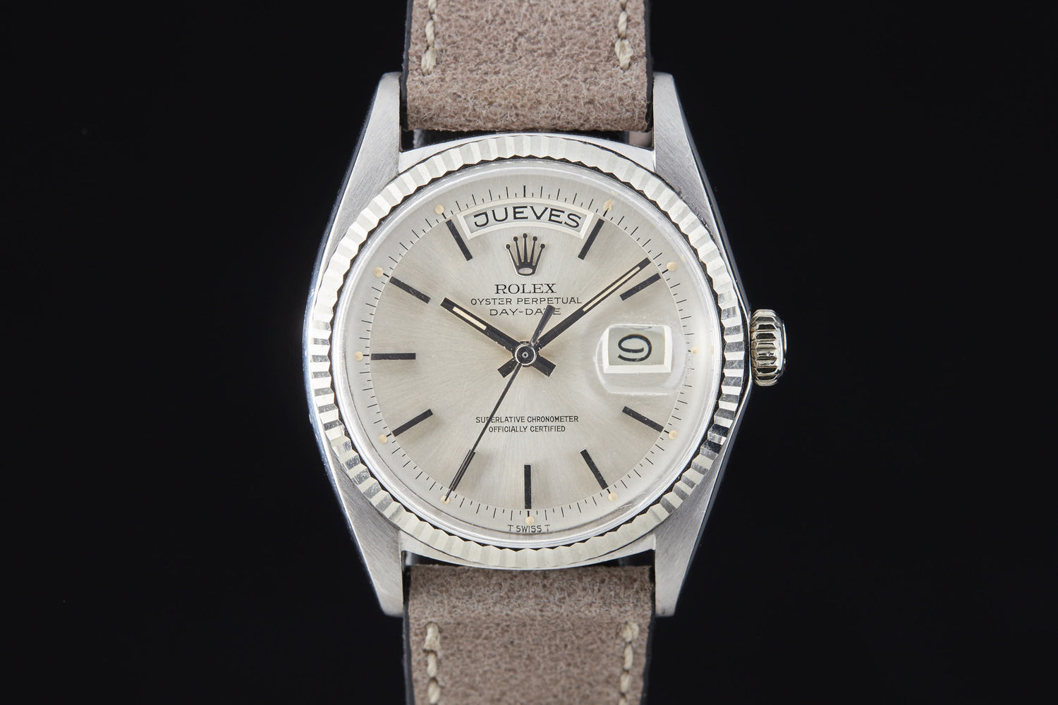 The Rolex Day Date by Rolex features a silver case, beige dial, Spanish day display ("JUEVES"), magnified date at 3 o’clock, brown leather strap, silver hour markers, automatic movement (Ref. 6511), shown on a black background.