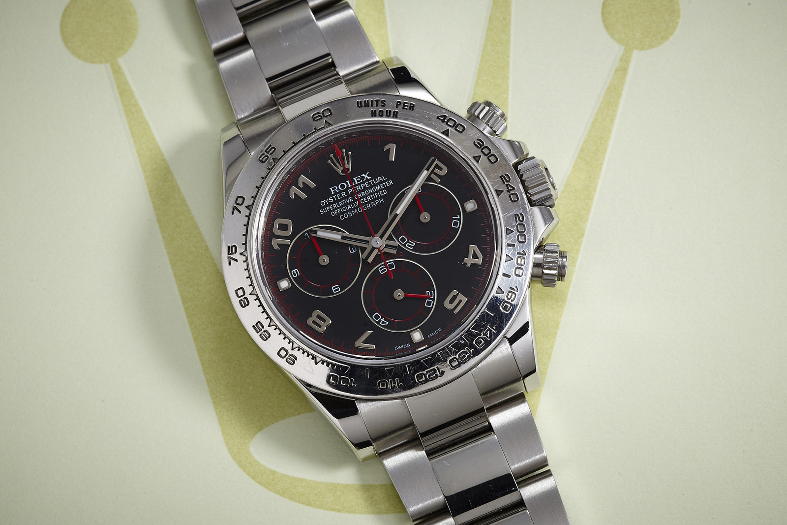 A Rolex Daytona Steel with Arabic Racing Dial, complete with box and papers, is shown on a backdrop featuring a large pale green Rolex crown logo.