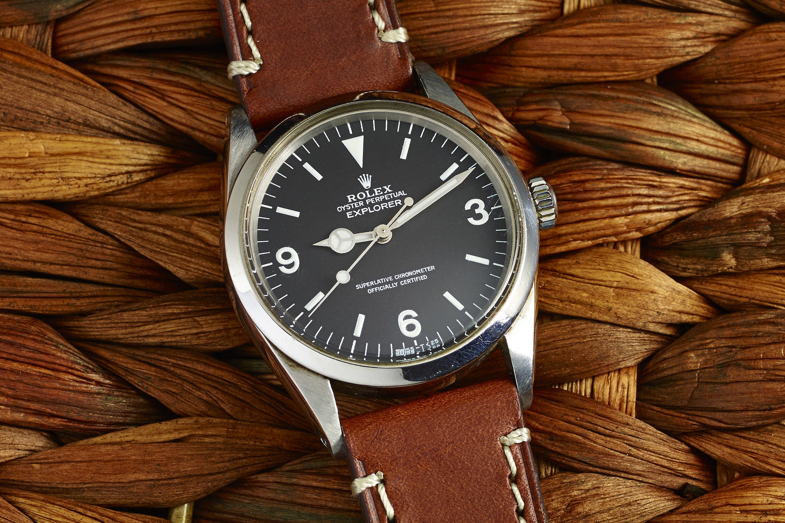A 1964 Rolex Explorer 1016 by Rolex, featuring a black dial, white hour markers, and a brown leather strap, rests on a natural woven surface—an iconic sports watch.