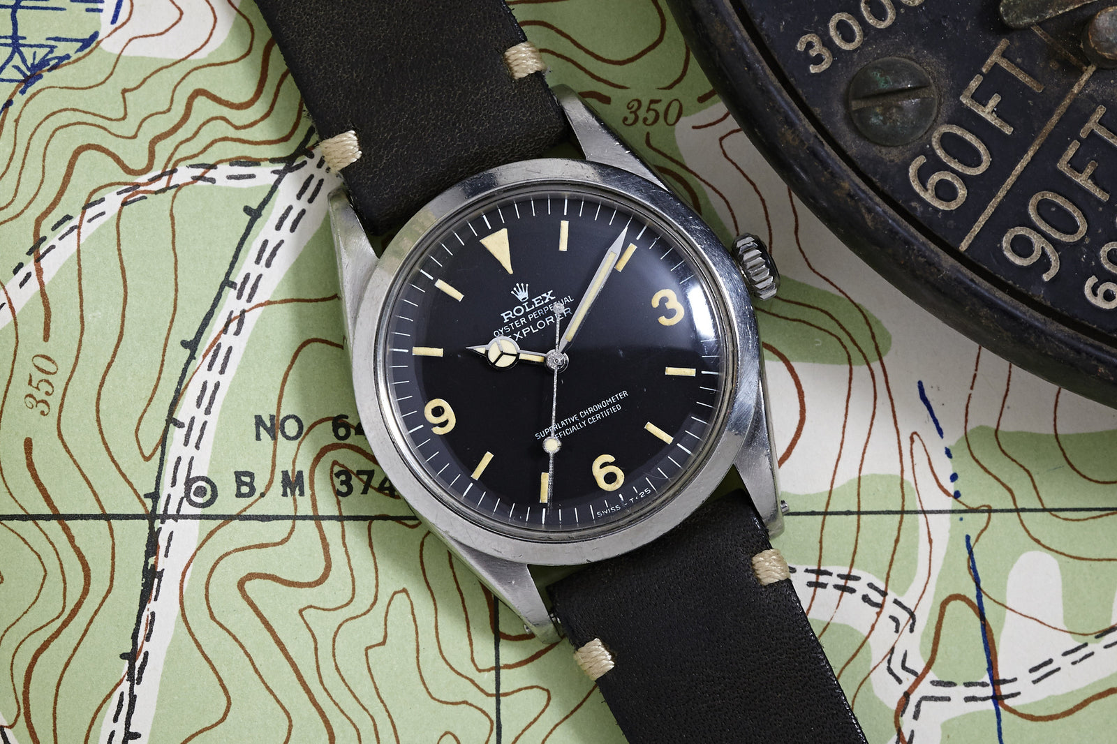 A Rolex Explorer 1016 - 1968 with a black dial and tan markers sits on a topographic map beside a round compass marked in feet.