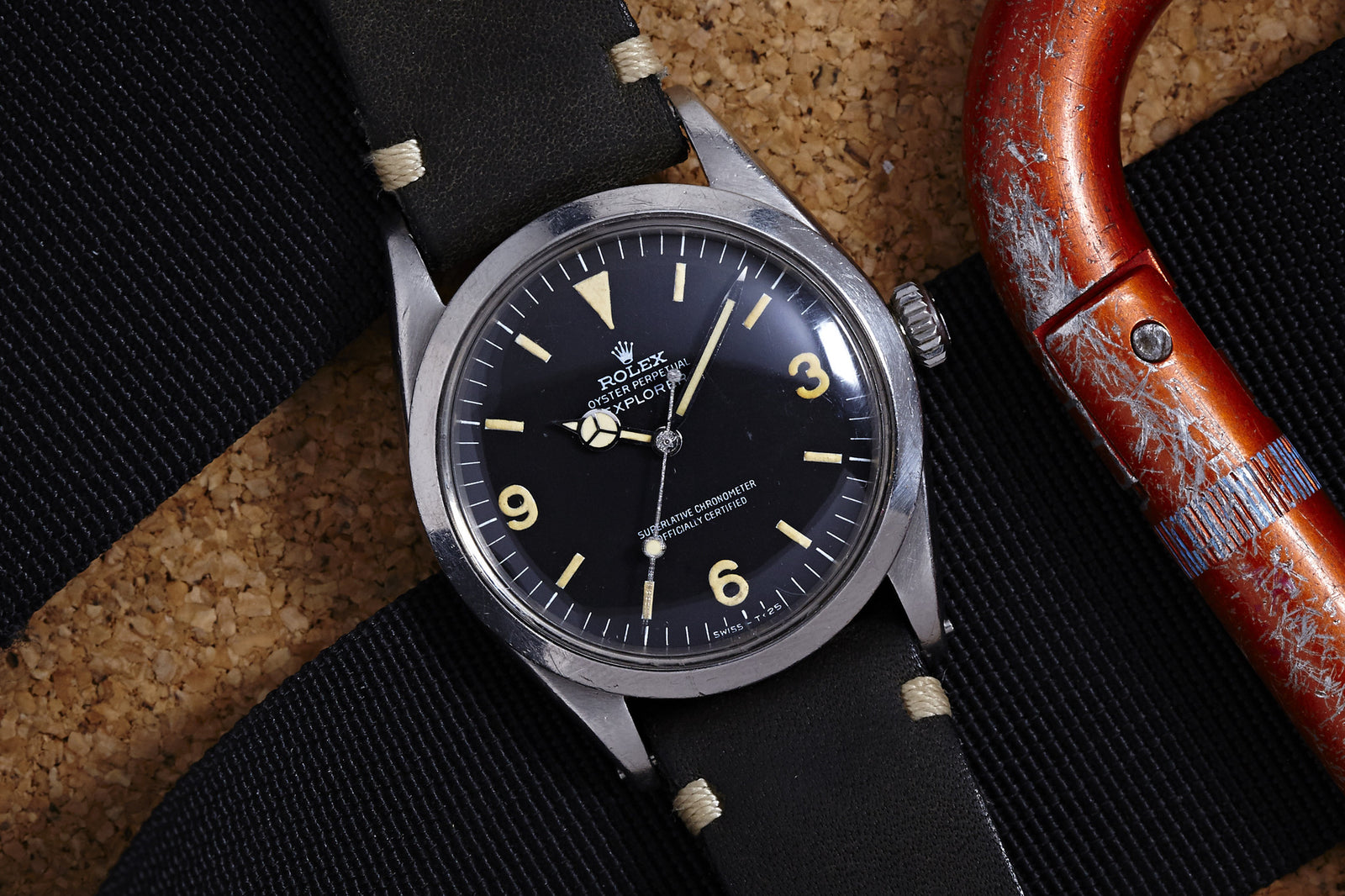 A Rolex Explorer 1016 - 1968 watch by Rolex, featuring a black dial and white hour markers with a black leather strap, is displayed on a textured surface beside a red climbing carabiner.