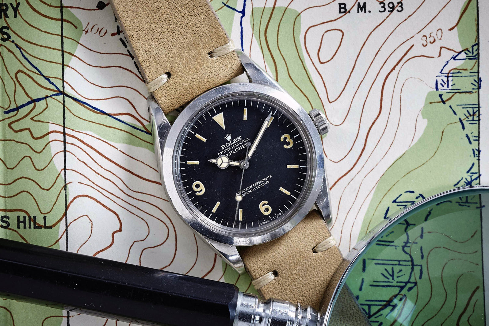 A Rolex Explorer by Rolex with a 3-6-9 black Arabic dial and tan leather strap rests on a topographic map, while a black magnifying glass handle is visible at the image’s lower left corner.