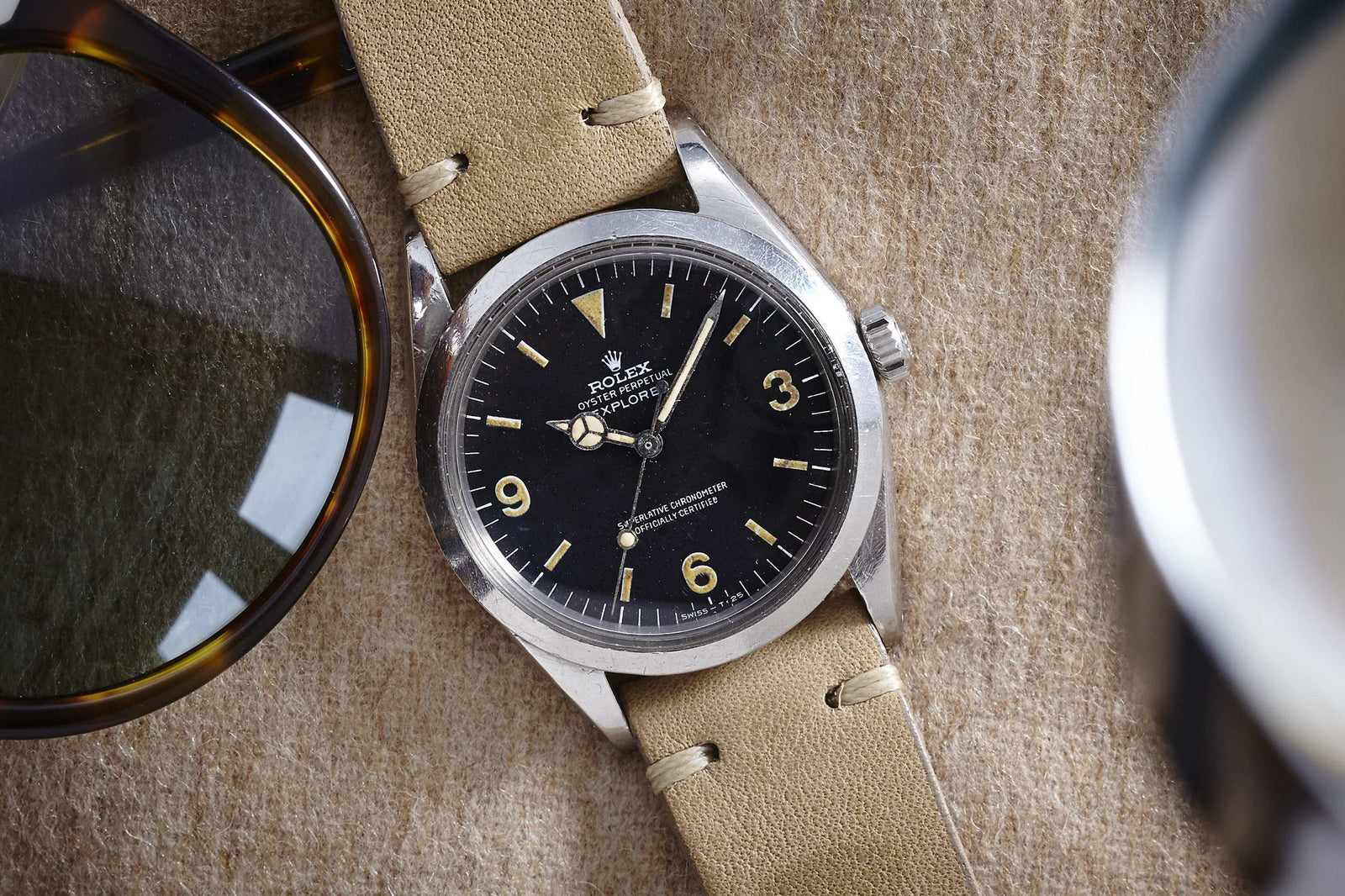 A Rolex Explorer watch with a black 3-6-9 Arabic dial and beige leather strap by Rolex rests on a textured tan surface next to sunglasses.