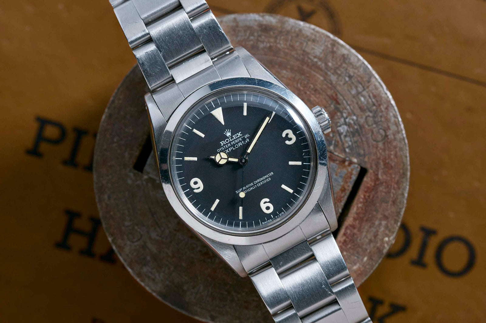 A Rolex Explorer by Rolex, featuring a stainless steel case, black dial with signature 3-6-9 markers, white hour markers, and silver hands, is displayed on a round metal surface against a brown background with partially visible black text.