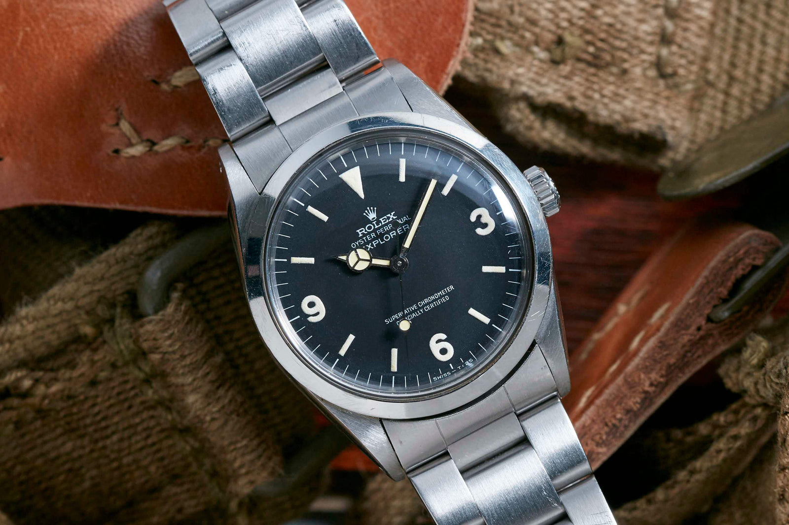 A close-up of the Rolex Explorer by Rolex, featuring the signature 3-6-9 dial with a black face, white hour markers, and stainless steel band, displayed on brown textured fabric.