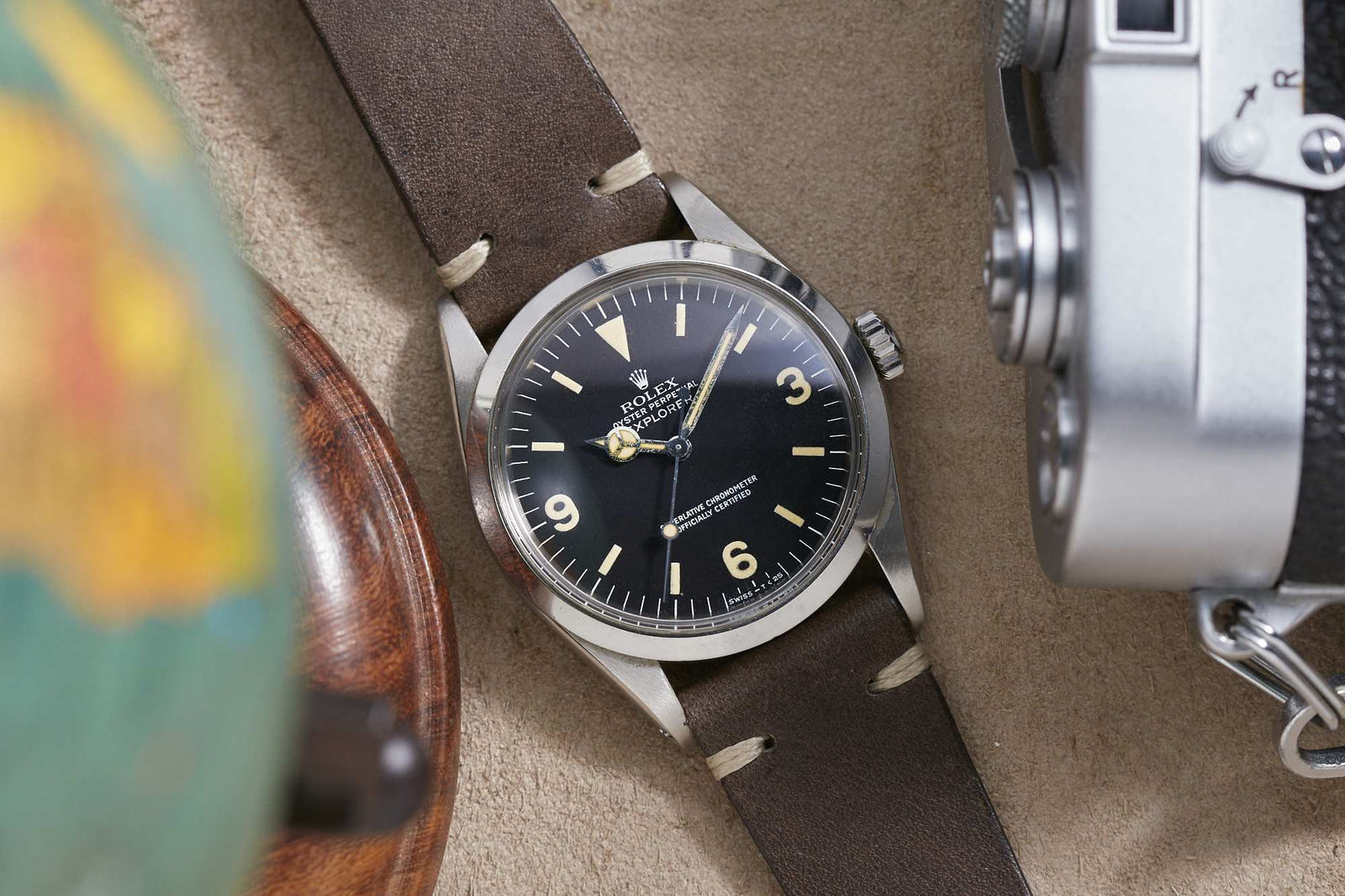 A Rolex Explorer 1016 wristwatch with a black dial and brown leather strap rests on a beige surface beside a globe and a silver camera. Detailed description coming soon!.