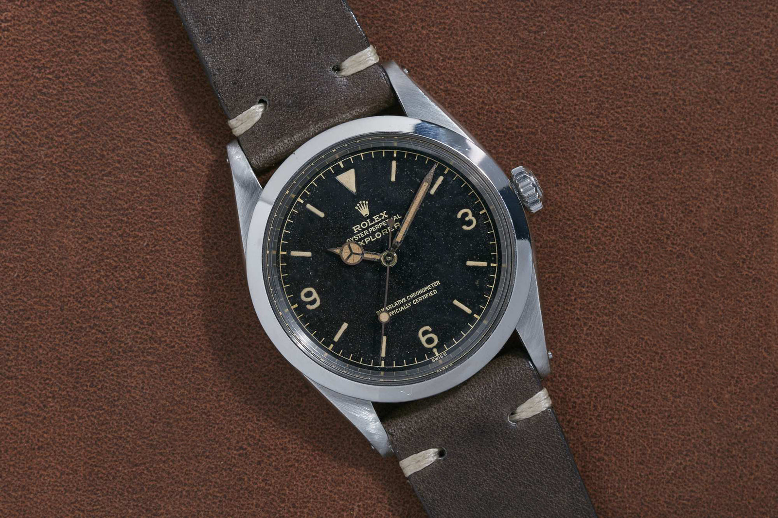 A Rolex Explorer 1016 wristwatch by Rolex featuring a black dial, cream hour markers and numerals, silver hands, and a brown leather strap, displayed on a textured brown surface. Detailed description coming soon!.