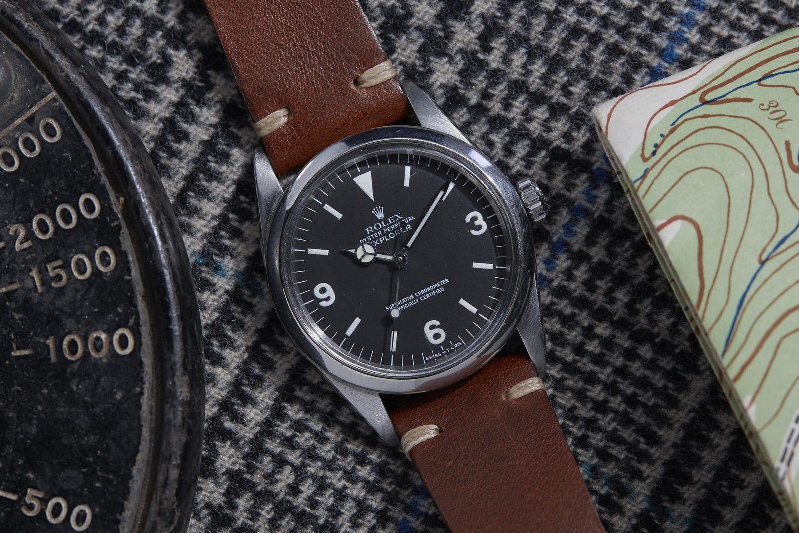 The Rolex Explorer 1016 by Rolex features a brown leather strap, black dial, and white numerals. Shown resting on houndstooth fabric beside a vintage gauge and a topographic map. Detailed Description Coming Soon!.