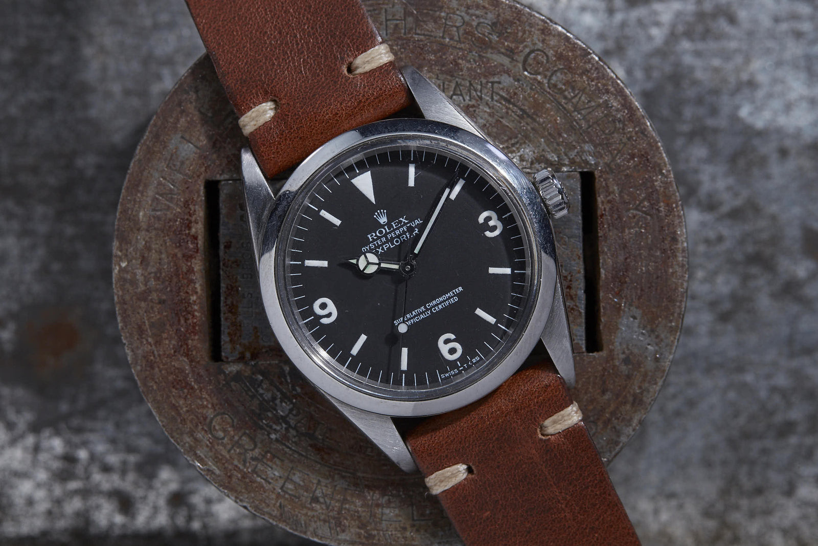 The Rolex Explorer 1016 by Rolex features a black dial, silver hour markers, and a brown leather strap, beautifully displayed on a round, weathered metal surface. Detailed description coming soon!.