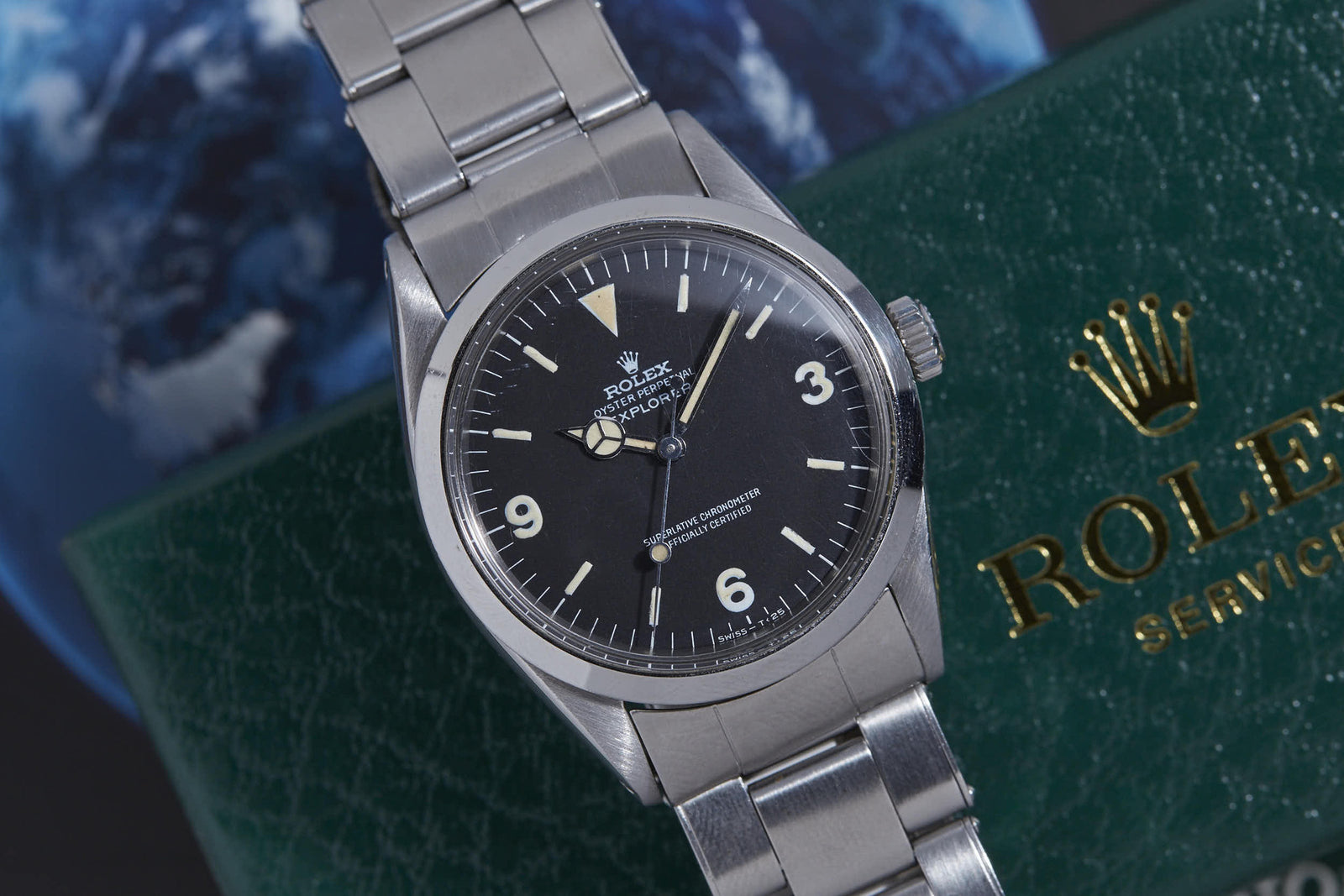 A close-up of the Rolex Explorer by Rolex, featuring a stainless steel case, black dial with white hour markers, and silver bracelet, set atop a green Rolex box with part of a globe visible in the background.