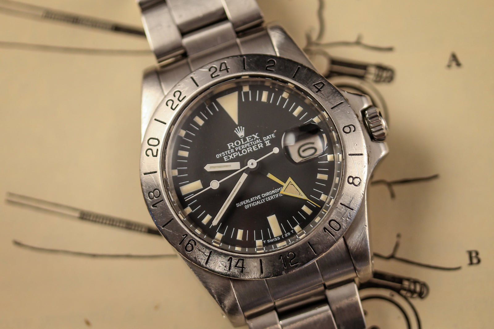 A close-up of the 1977 Rolex Explorer II 1655 by Rolex, featuring a brushed stainless steel band, 24-hour bezel, black dial with date window, and yellow 24-hour hand, resting on a sepia map marked "A" and "B".