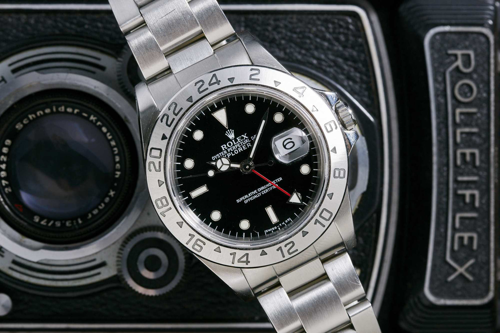 A Rolex Explorer II by Rolex with a 24-hour bezel and black dial sits atop a vintage Rolleiflex camera, showcasing the camera's visible lens and branding in the background.