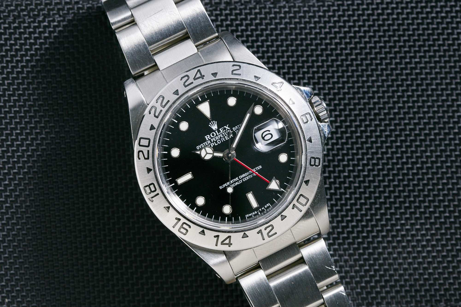 The Rolex Explorer II by Rolex features a stainless steel case, black dial, silver hour markers, date window at 3 o’clock, 24-hour bezel, and metal link bracelet. It's displayed on a textured dark surface.