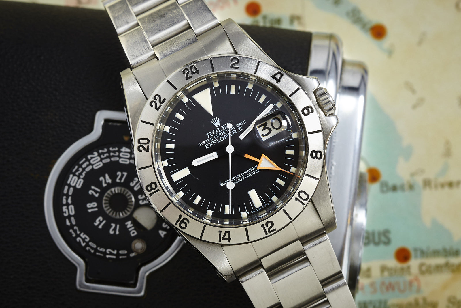 A Rolex Explorer II speleology watch by Rolex with a stainless steel band sits on a black camera case, map in the background. The watch features time, date, an orange 24-hour hand, and the signature 24-hour bezel.