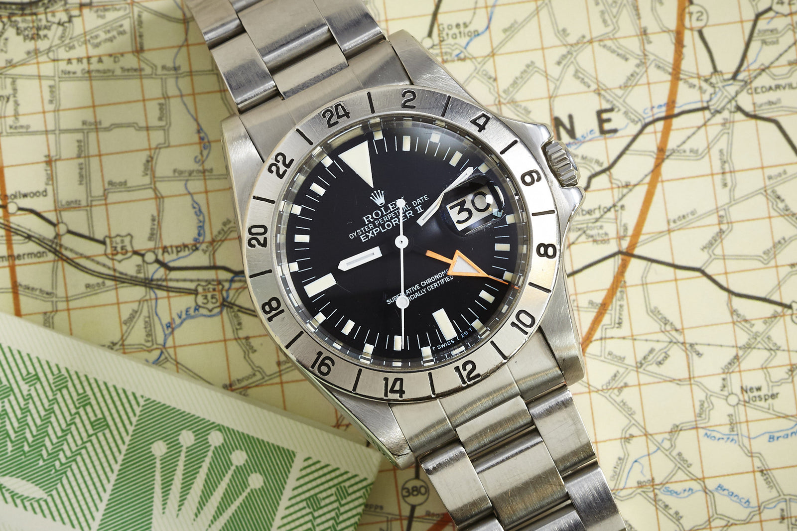 A Rolex Explorer II watch with a black dial, date display, and 24-hour bezel by Rolex rests on a detailed map. A portion of a green and white Rolex booklet appears in the lower left corner.