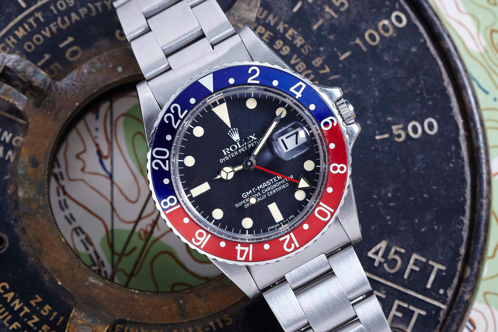 A Rolex GMT-Master wristwatch by Rolex with a red and blue bezel, black dial, and silver metal bracelet is displayed on a vintage map alongside a compass with elevation markings.