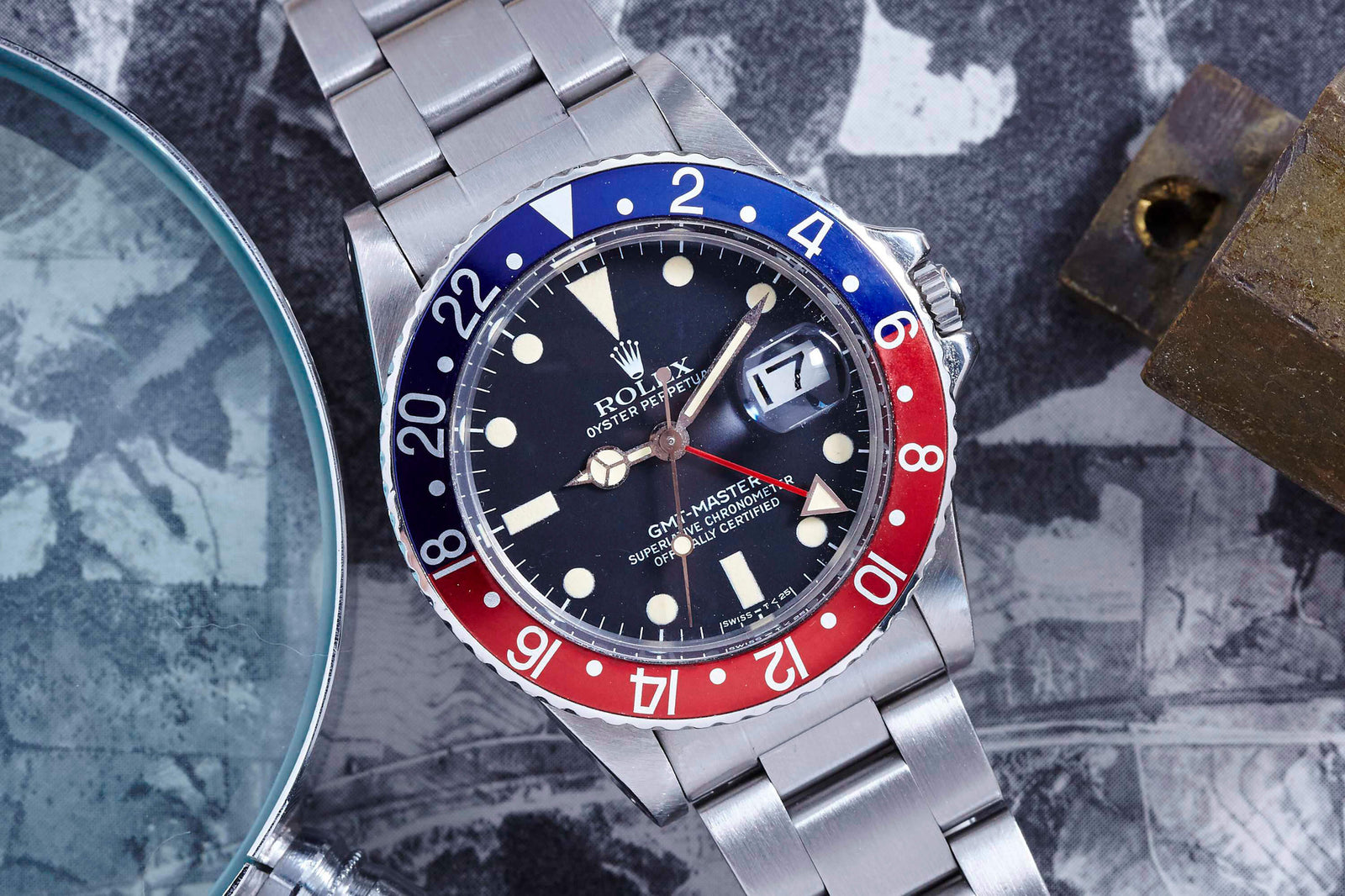 A close-up shows the Rolex GMT-Master by Rolex, featuring a red and blue bezel and black dial, displayed beside a magnifying glass and padlock on a textured surface.